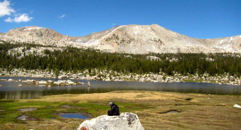 A person sits on a rock near a body of water. On the other side of water is a rocky shore, evergreen trees and a mountain. 